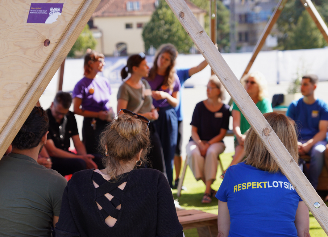 Foto von Menschen die in einem Veranstalltungszelt zusammensitzen. Eine Person trägt ein T-shirt auf dem Respektlotse steht.